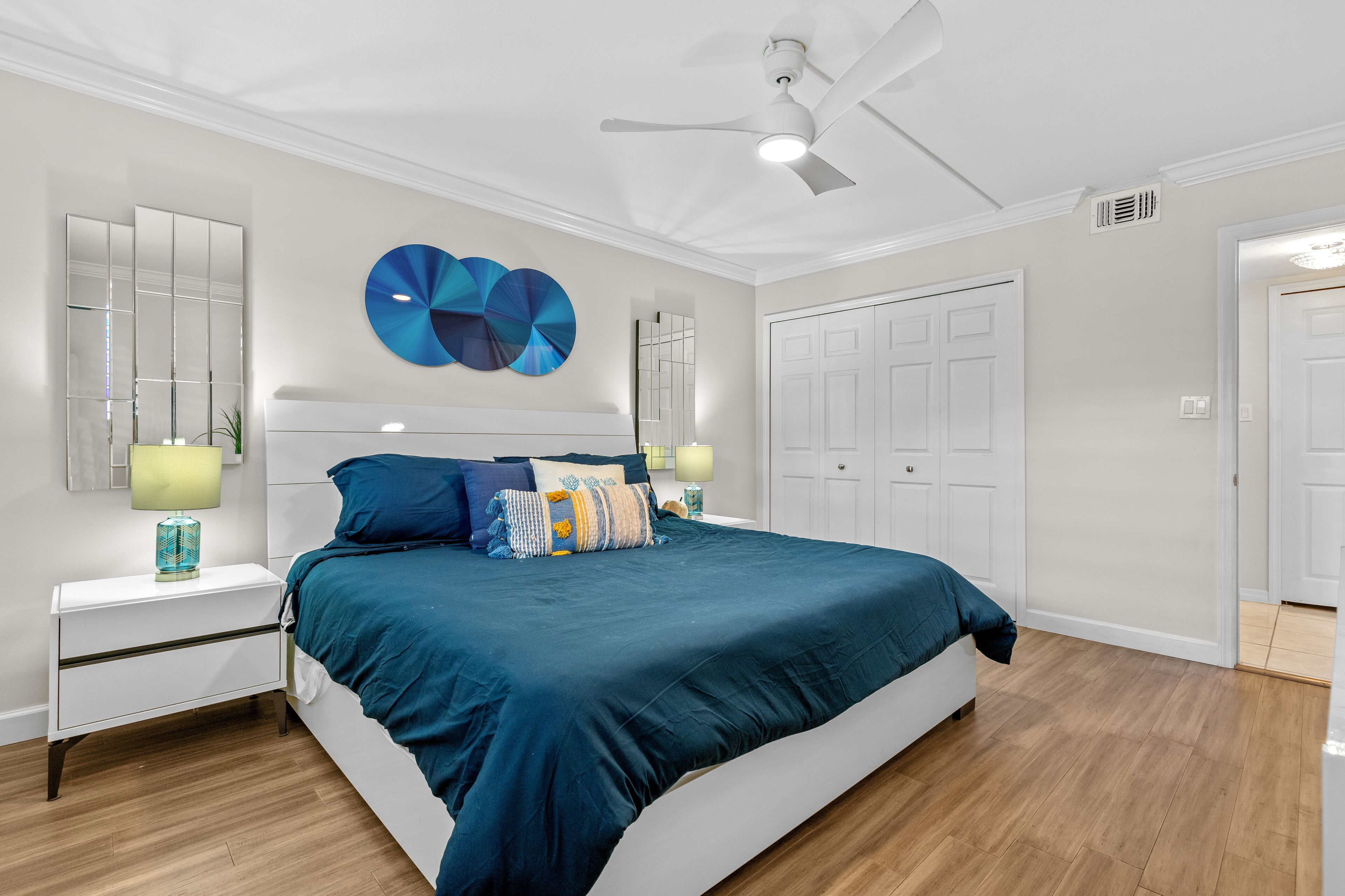 A bedroom with a large bed, a ceiling fan, a nightstand with a lamp, and a wall-mounted mirror.