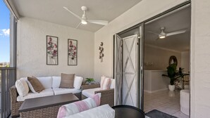 Een balkon met een witte bank, een klein tafeltje en een stoel. Er zijn plafondventilatoren en decoratieve wandkunst.