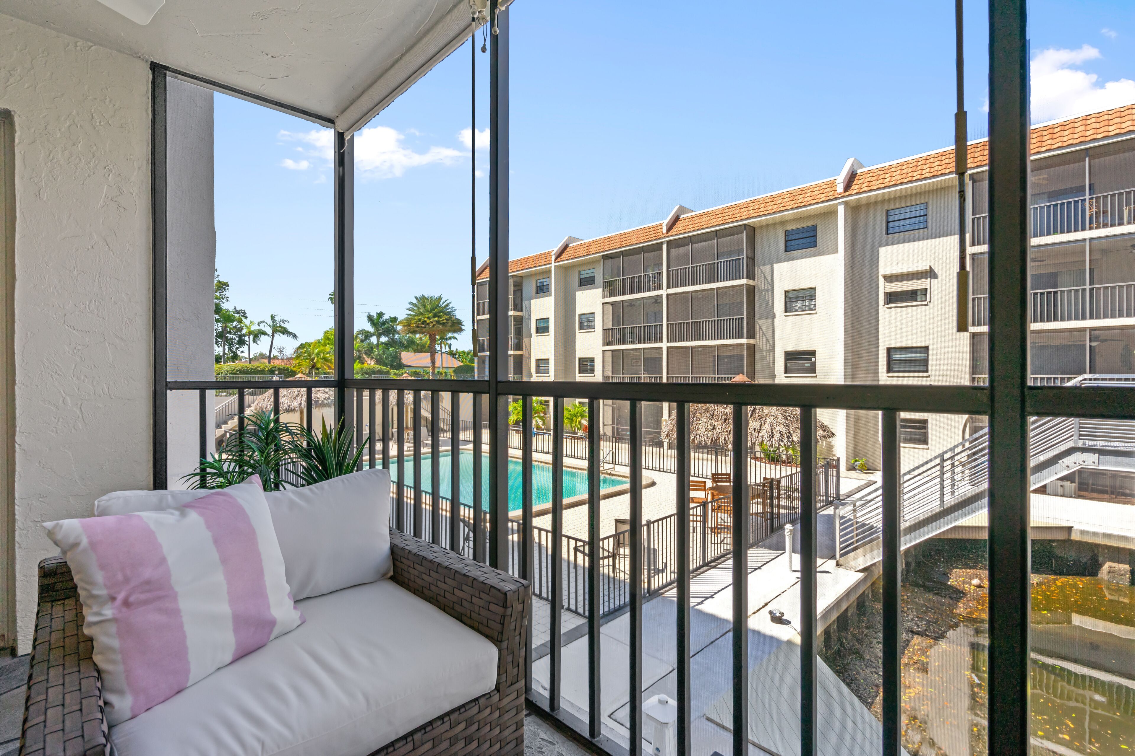 A balcony with a wicker sofa and a view of a swimming pool and adjacent buildings.