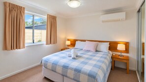 A hotel room with a bed, two bedside tables, a window with curtains, a wall-mounted air conditioner, and a ceiling light. - Cape View TwentyTwo – a modern 2-bedroom, 2-bathroom villa (Broadwater)