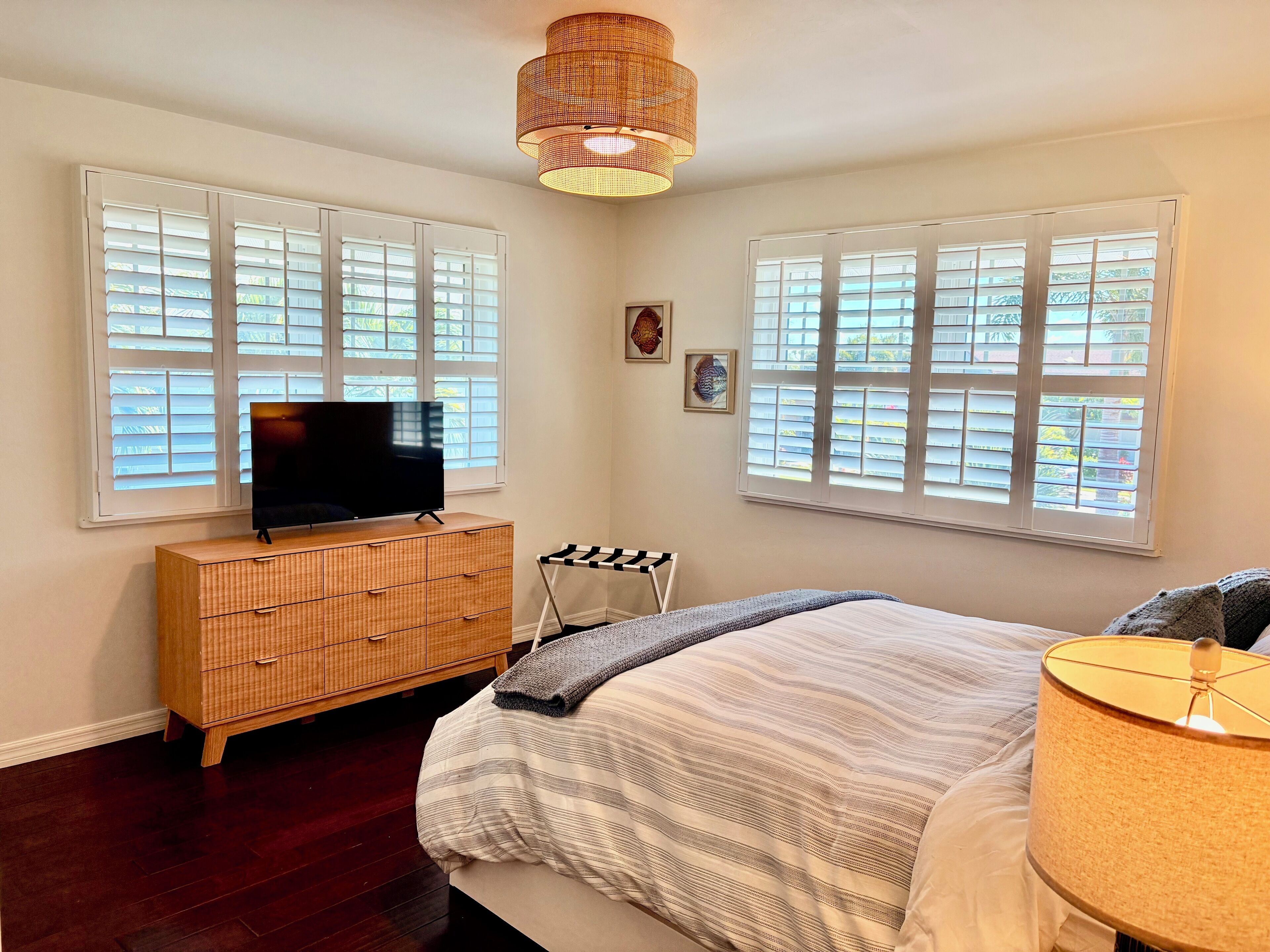 A bedroom with a bed, a TV on a wooden dresser, and two windows with blinds.