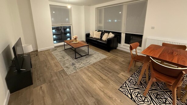 Un salon moderne avec un canapé en cuir noir, une table basse en bois et une table à manger avec des chaises. On y trouve un téléviseur posé sur un meuble noir, une grande fenêtre et une cheminée.