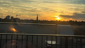 Une vue sur le coucher de soleil depuis une terrasse sur le toit, avec une balustrade, des galets et le profil d’une ville lointaine.