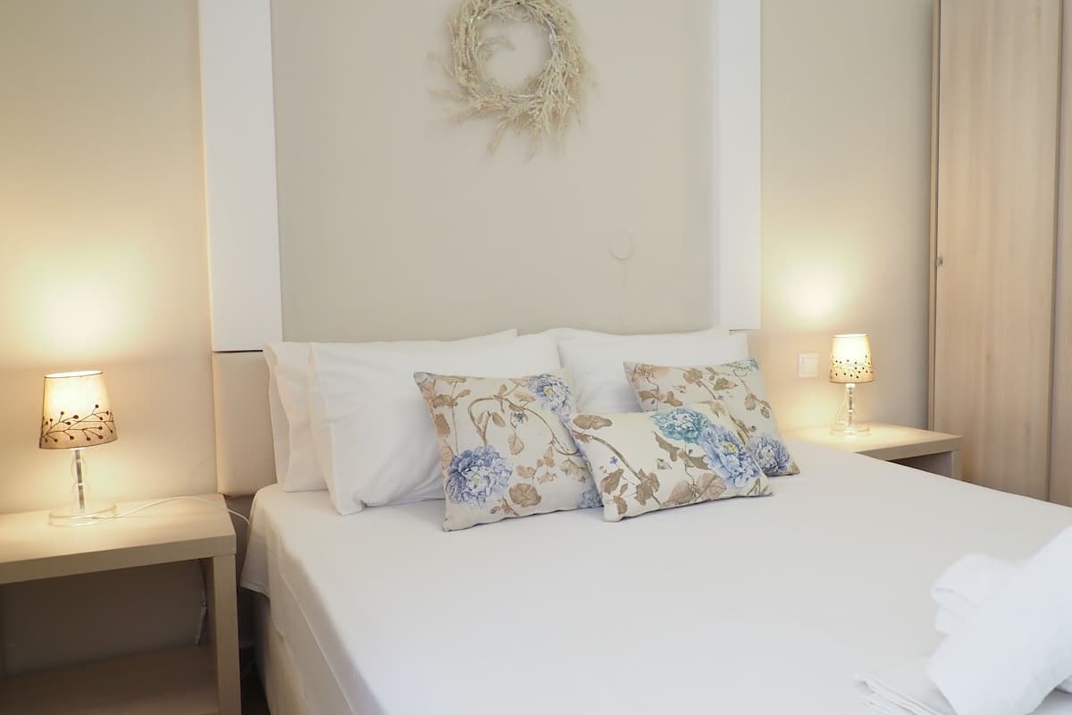 A neatly made bed with floral pillows, two bedside lamps, a wooden wardrobe, and a small table with towels.