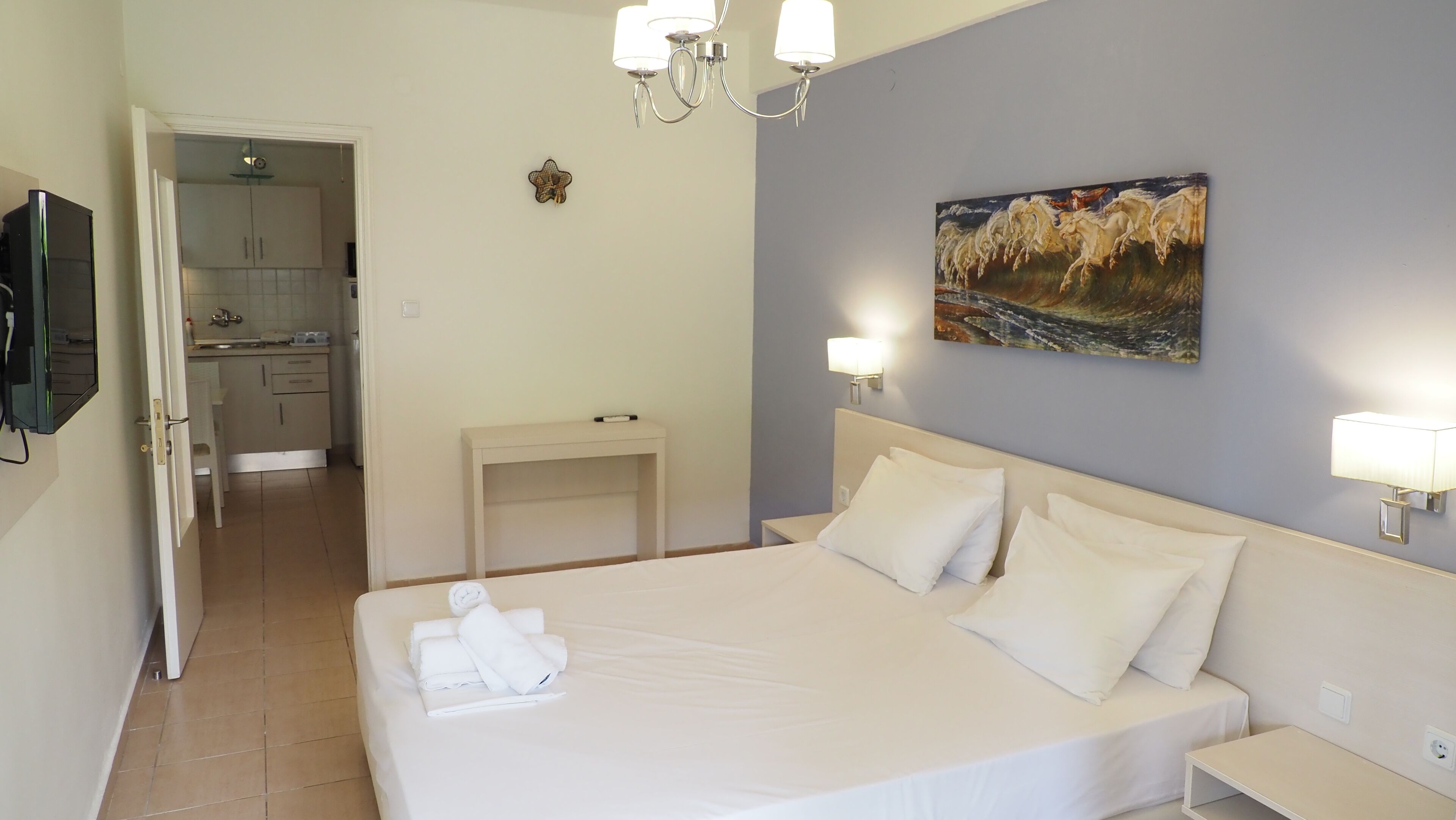 A hotel room with a bed, bedside tables, a painting, and a view into the kitchen.