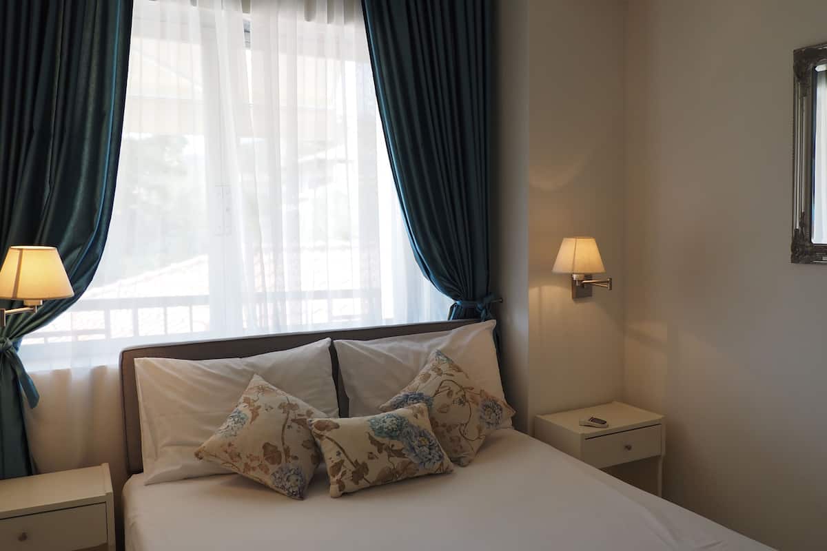 A hotel room with a bed, two bedside lamps, a nightstand, a mirror, and a window with curtains.