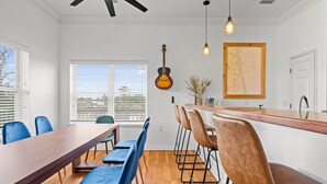 A dining area with a long wooden table, blue chairs, a bar counter with stools, a guitar, and a map on the wall. - Cedar Island Coastal Retreat | Gulf Access, Huge Deck & Fami (Taylor County)