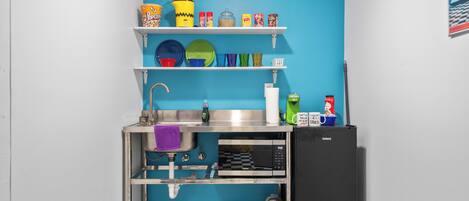 A compact kitchen with a blue wall, a black and white checkered floor, a sink, a microwave, a refrigerator, and shelves with various kitchen items.