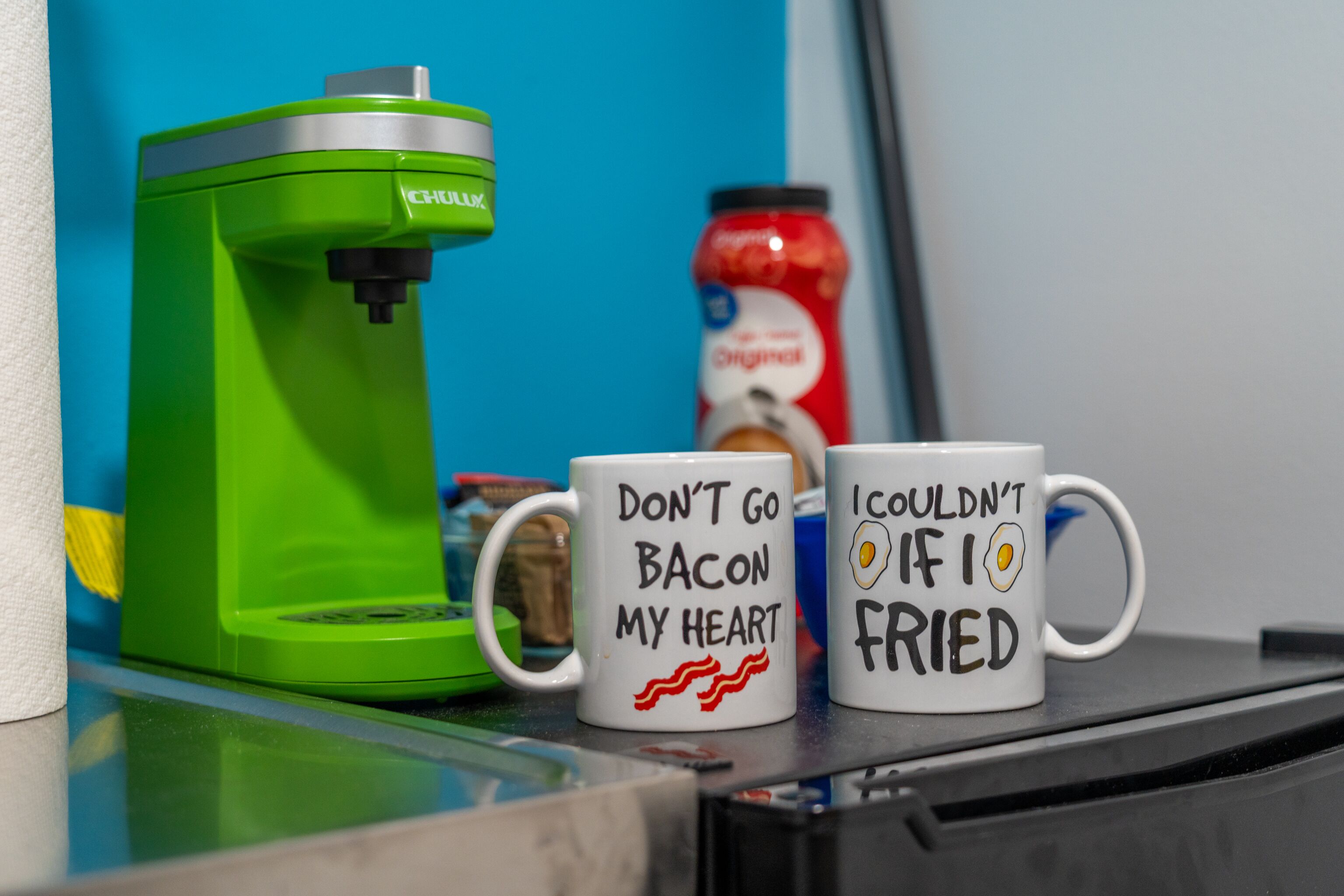 Two mugs with humorous text, a green coffee maker, and a jar of ketchup on a countertop.