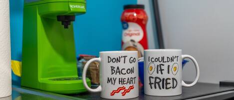 Two mugs with humorous text, a green coffee maker, and a jar of ketchup on a countertop.