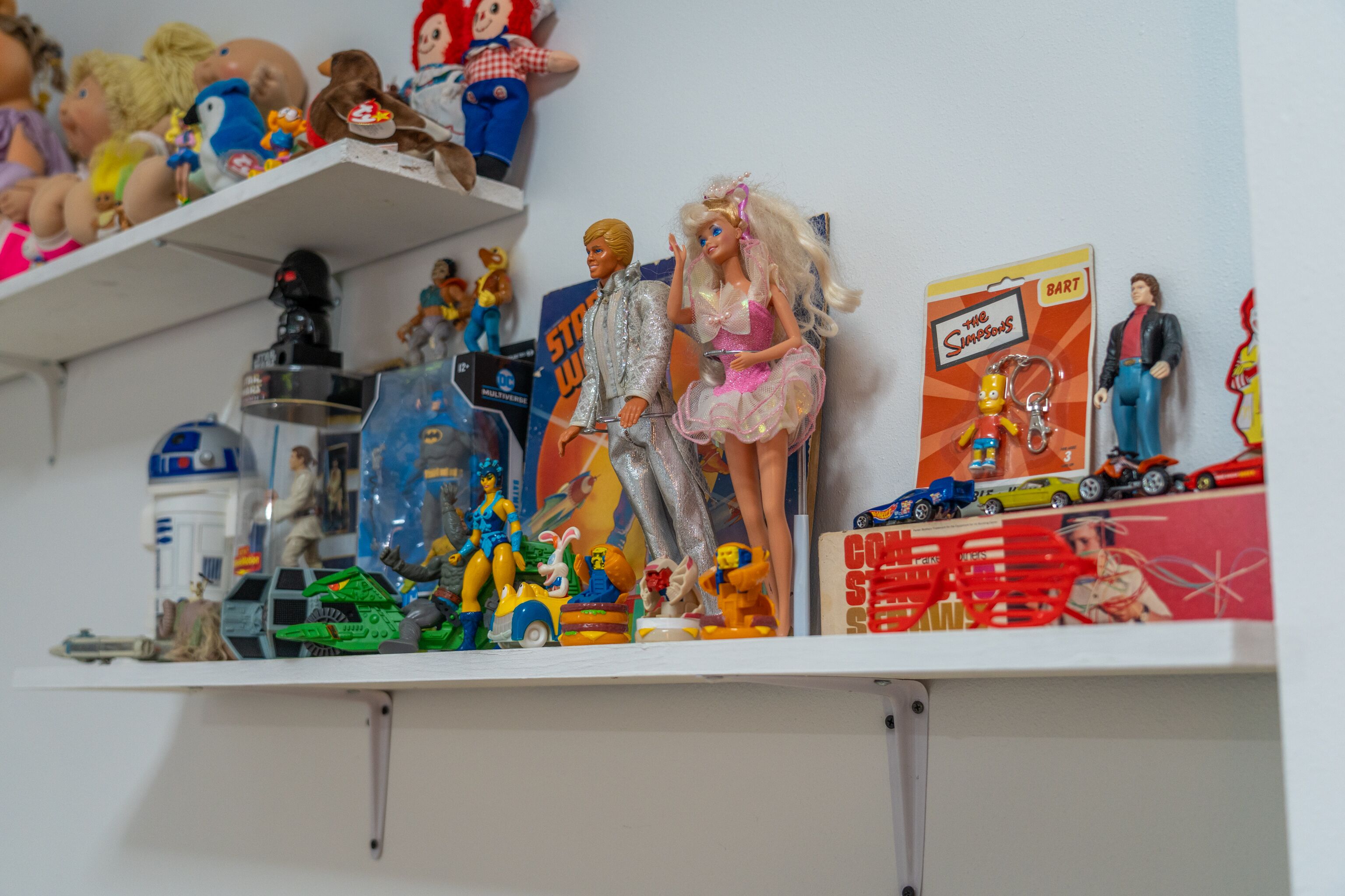 Una estantería con diversos objetos de colección, incluyendo figuras de acción, muñecas y coches de juguete.