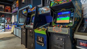 Een rij spelautomaten met verschillende spellen, waaronder 'Space Invaders' en 'Pac-Man', in een ruimte met een betonnen vloer en muren versierd met posters.