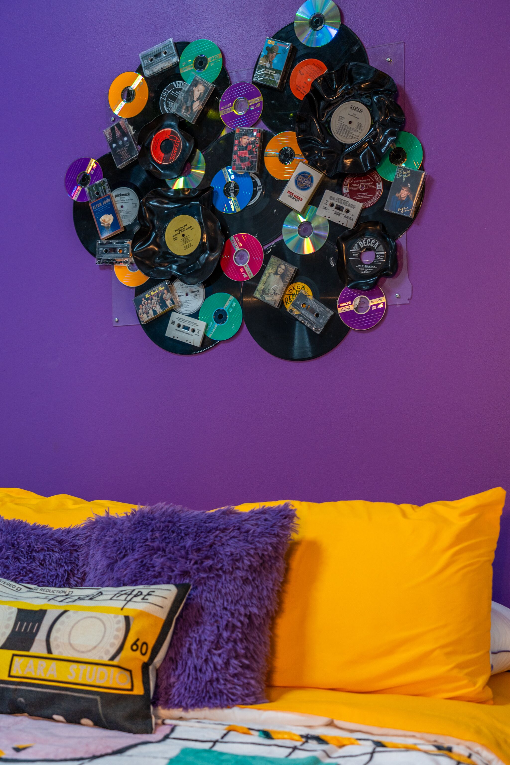 Una cama con una manta morada y amarilla, un almohadón esponjoso morado, y una pared decorada con varios CDs y discos de vinilo.