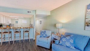 A living room with a blue sofa and matching chairs, a coffee table, and a kitchen area with bar stools.