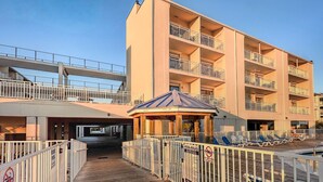 A wooden boardwalk leading to a pool area with lounge chairs, a gazebo, and a multi-story building with balconies.