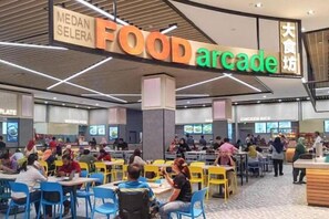 Un patio de comidas con múltiples mesas y sillas, y carteles que señalan distintas opciones de comida.