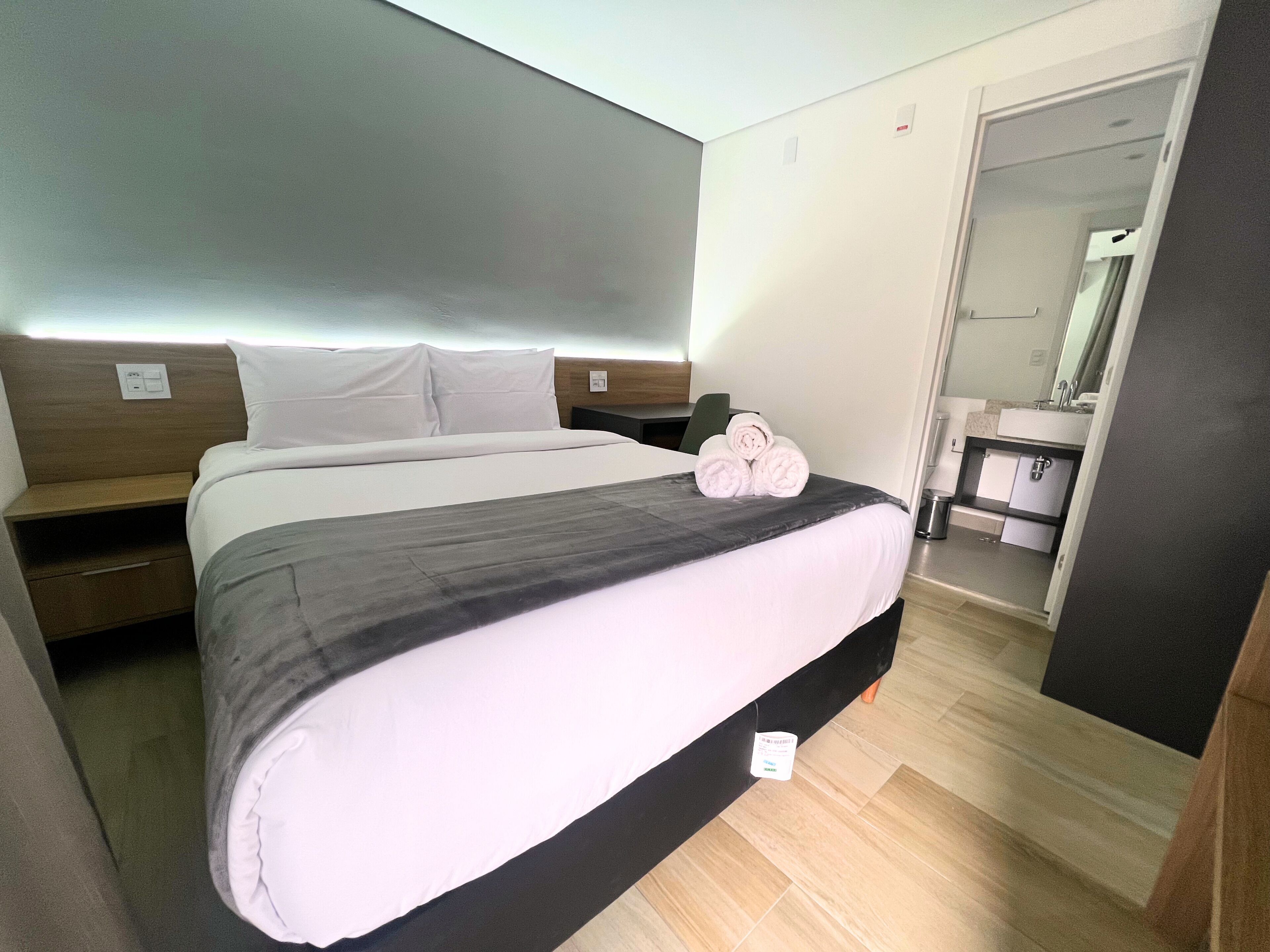 Una camera d'albergo con un letto, un comodino e un bagno visibile attraverso una porta aperta.