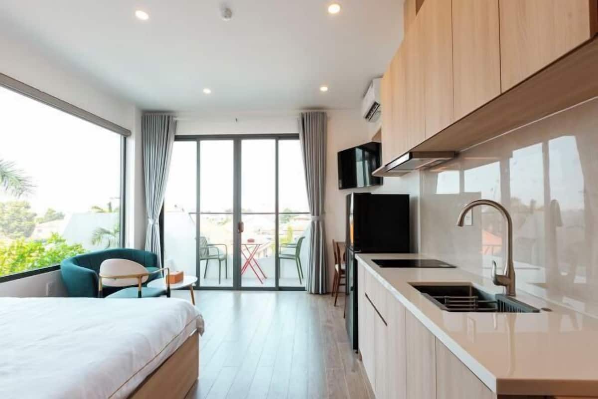A modern hotel room with a bed, a kitchenette, a TV, and a balcony with a table and chairs.