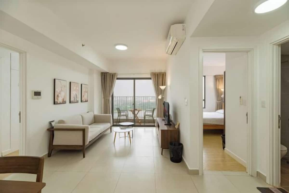 A modern living room with a sofa, a coffee table, and a TV stand. There is a balcony with a view, and an air conditioning unit on the wall.