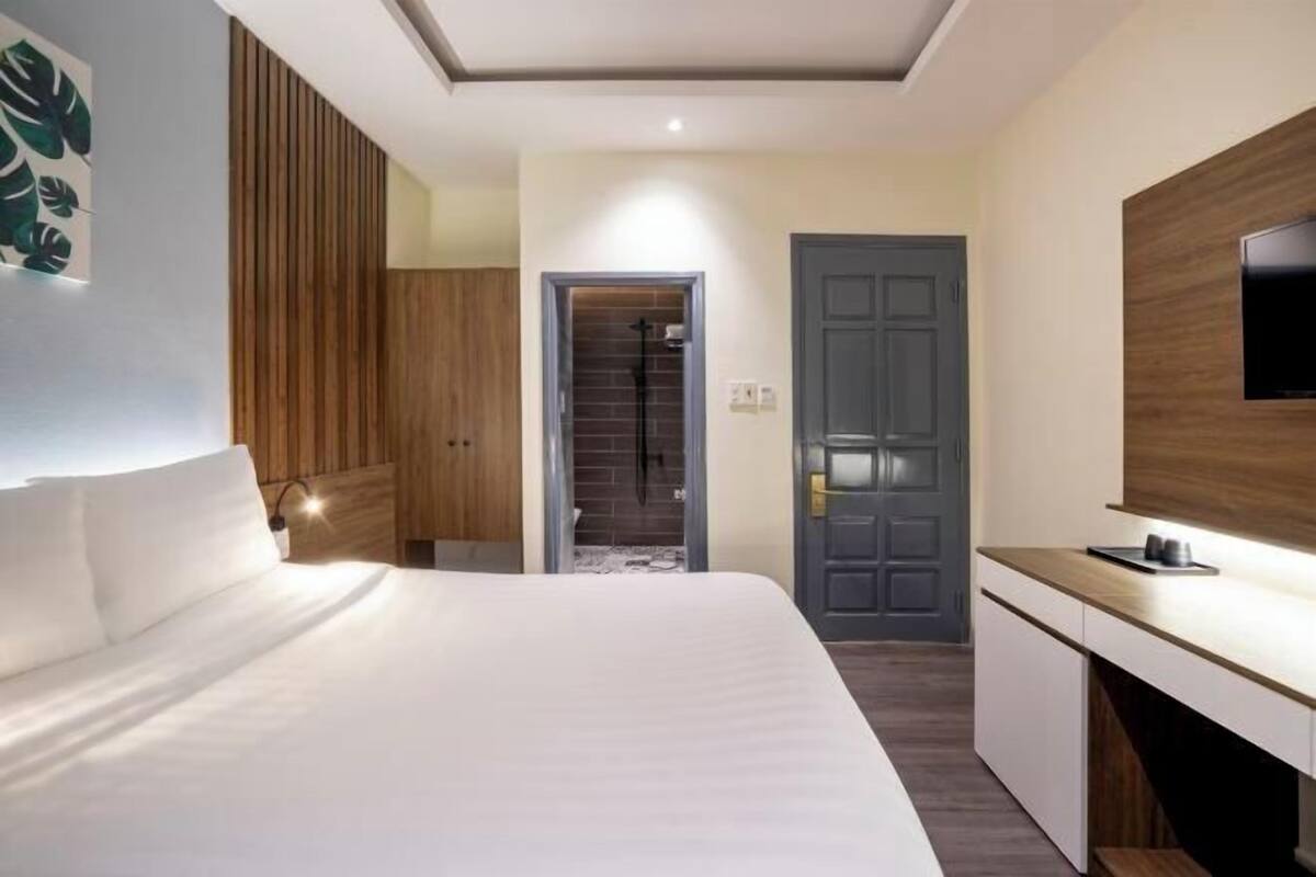 A modern hotel room with a large bed, a wooden wardrobe, a desk with a flat-screen TV, and a shower area.