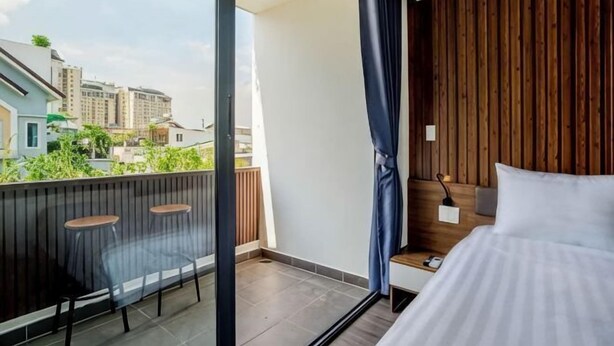 Ein Schlafzimmer mit Bett, Nachttisch, einem Fenster mit Vorhängen, einem Balkon mit Stühlen und Blick auf Gebäude.
