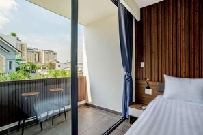 Un dormitorio con cama, mesita de noche, ventana con cortinas, balcón con sillas y vistas a edificios.