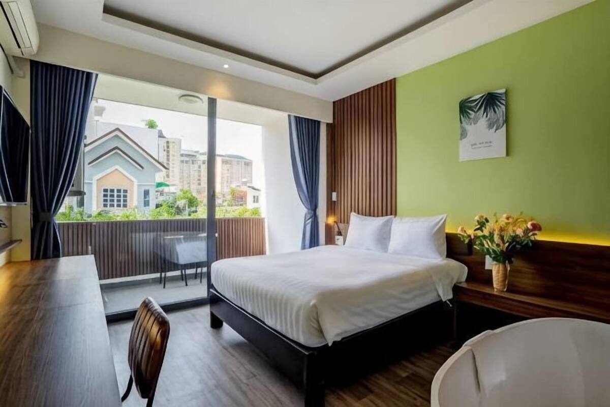 A hotel room with a bed, a desk, a chair, a window with curtains, and a view of buildings outside.