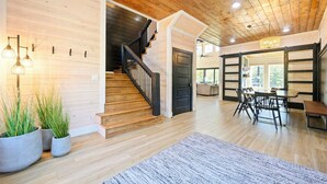 A spacious living area with a wooden staircase, a dining table with chairs, and a large window providing natural light. - Luxury 4BR Cabin on Pond for 18p- Pool, Sauna, Hot Tub & Game Room in Broken Bow (Broken Bow)