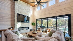 A modern living room with a stone fireplace, large windows, and a ceiling fan. - Luxury 4BR Cabin on Pond for 18p- Pool, Sauna, Hot Tub & Game Room in Broken Bow (Broken Bow)