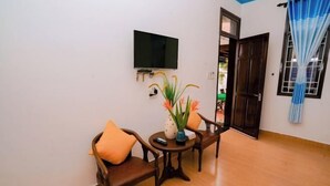 A room with a TV mounted on the wall, a small round table with a vase of flowers, two wooden chairs, and a wooden floor. - Tra Que Green Villa Hoi An (Hoi An)