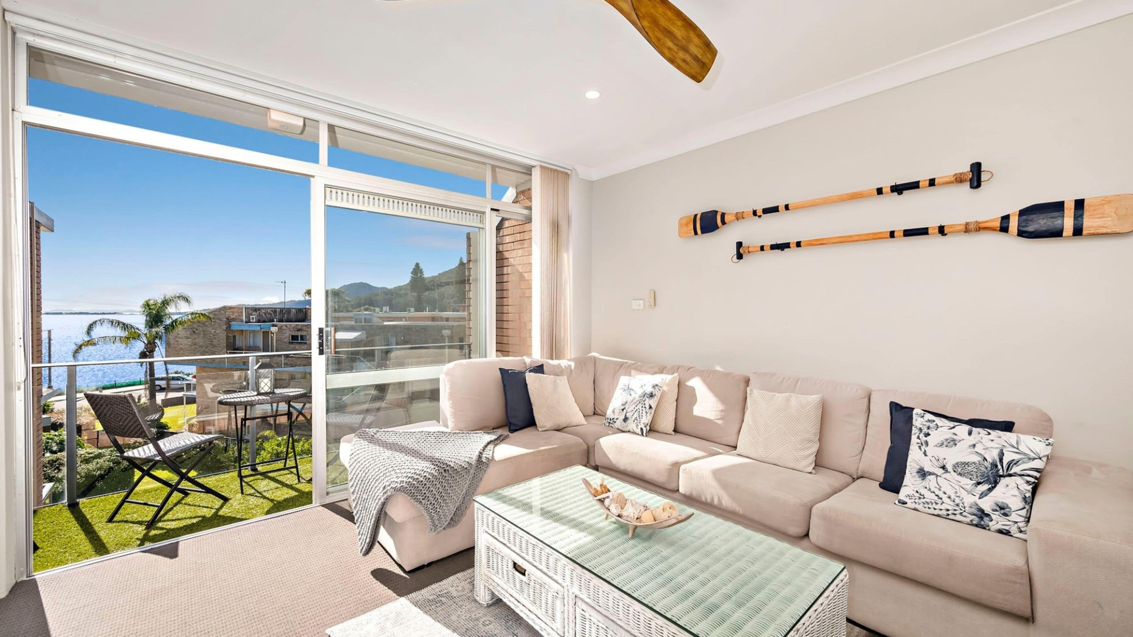 Een moderne woonkamer met een grote glazen deur die leidt naar een balkon, een beige bank en twee houten paddles aan de muur.