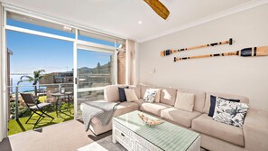 Een moderne woonkamer met een grote glazen deur die leidt naar een balkon, een beige bank en twee houten paddles aan de muur.