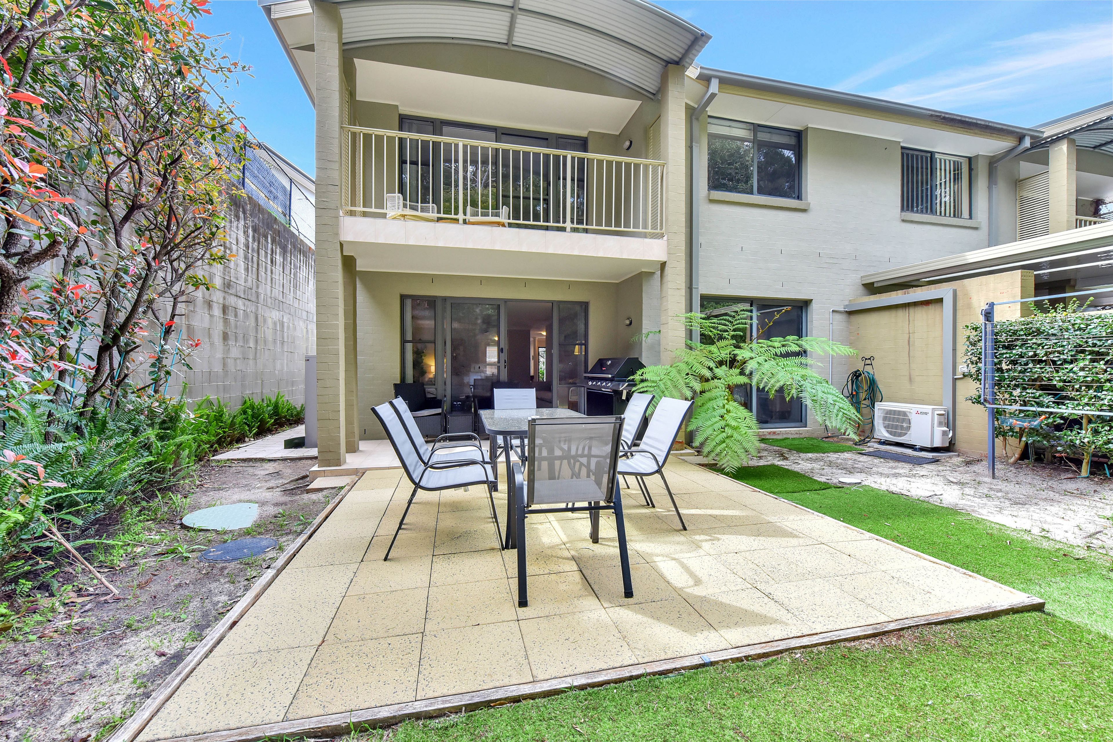A two-story house with a patio area featuring a table and chairs, a barbecue grill, and a small garden with ferns.