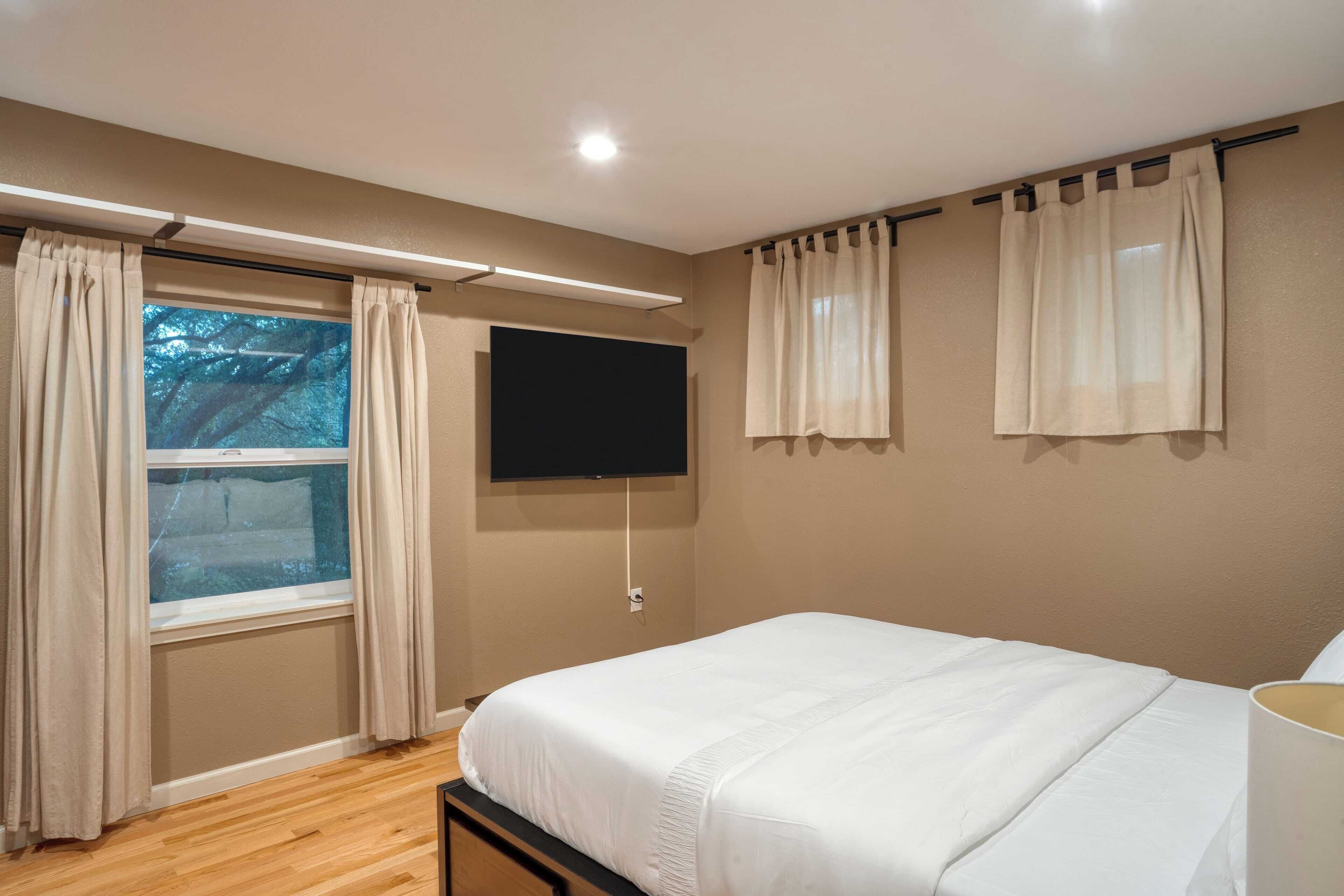 Un dormitorio con cama, ventana con cortinas, televisión de pantalla plana en la pared y una lámpara en el techo.