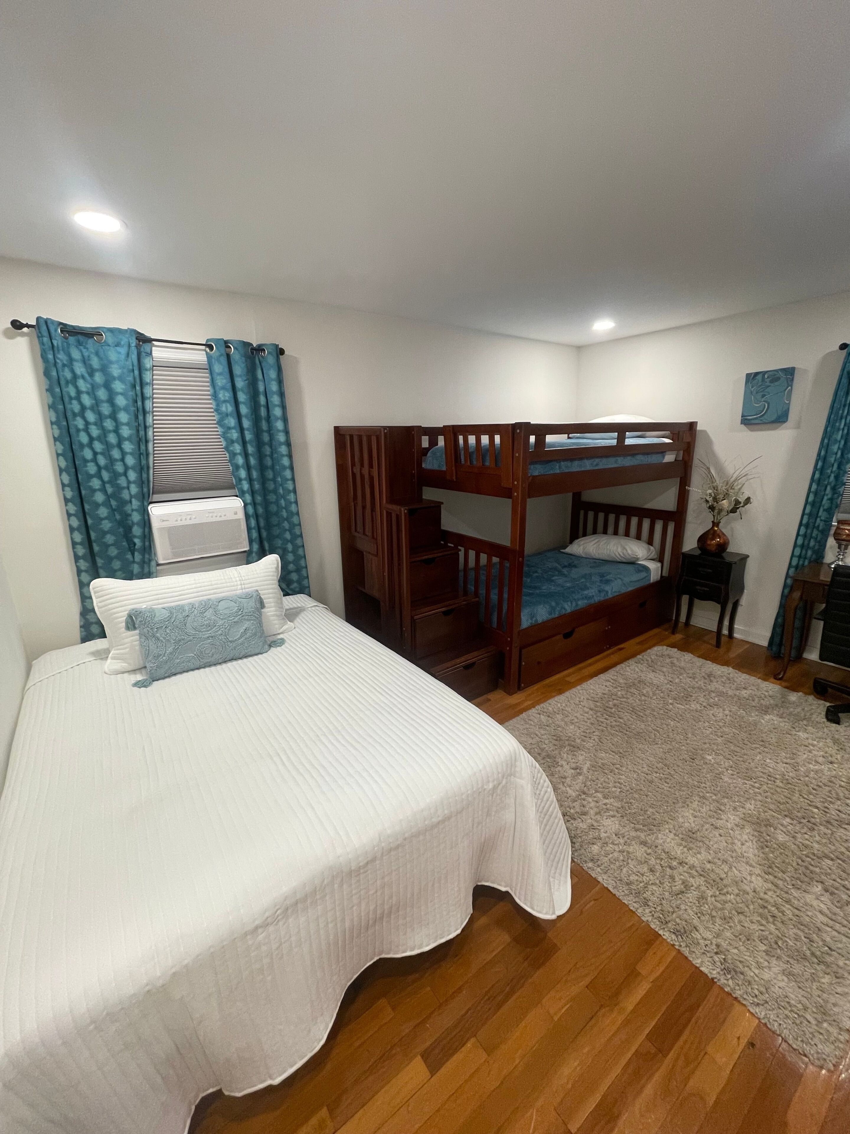 A bedroom with a bunk bed, a single bed, a wooden floor, a rug, a desk, and a chair.