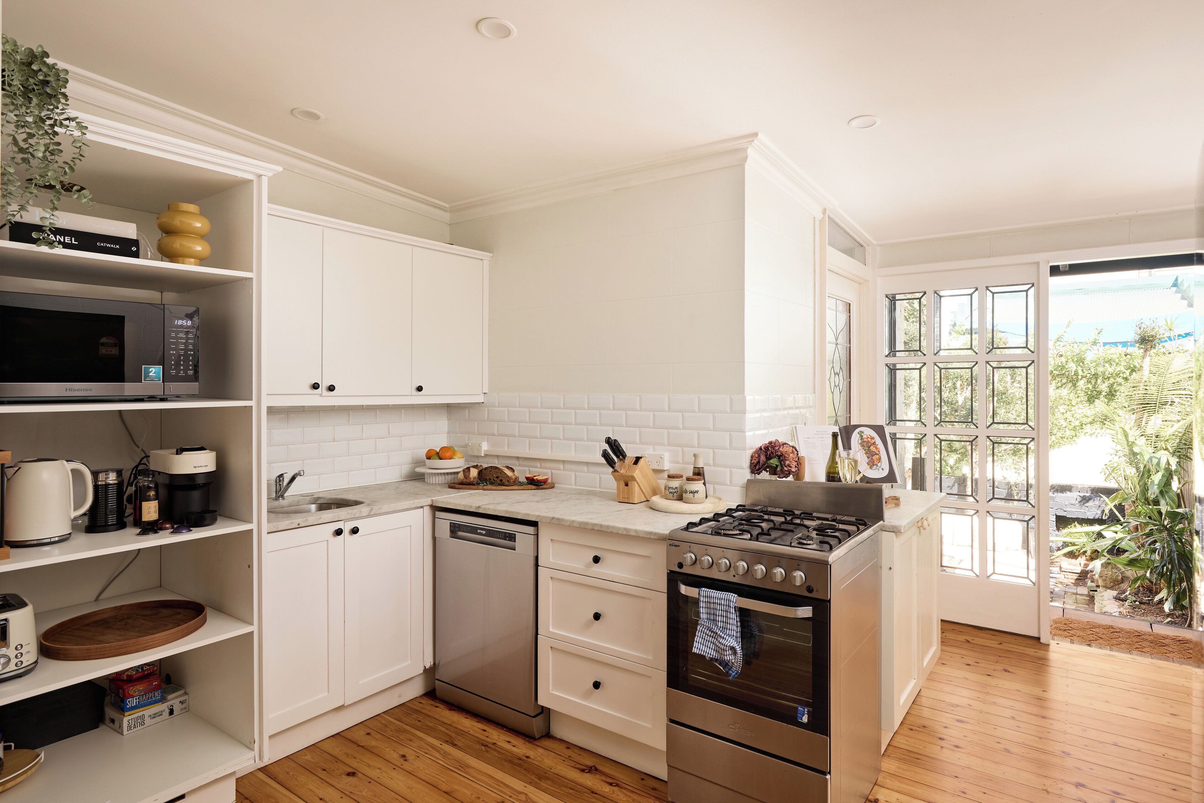 A modern kitchen with white cabinets, a dishwasher, a stove, and a sink. There is a window with a view of greenery outside.