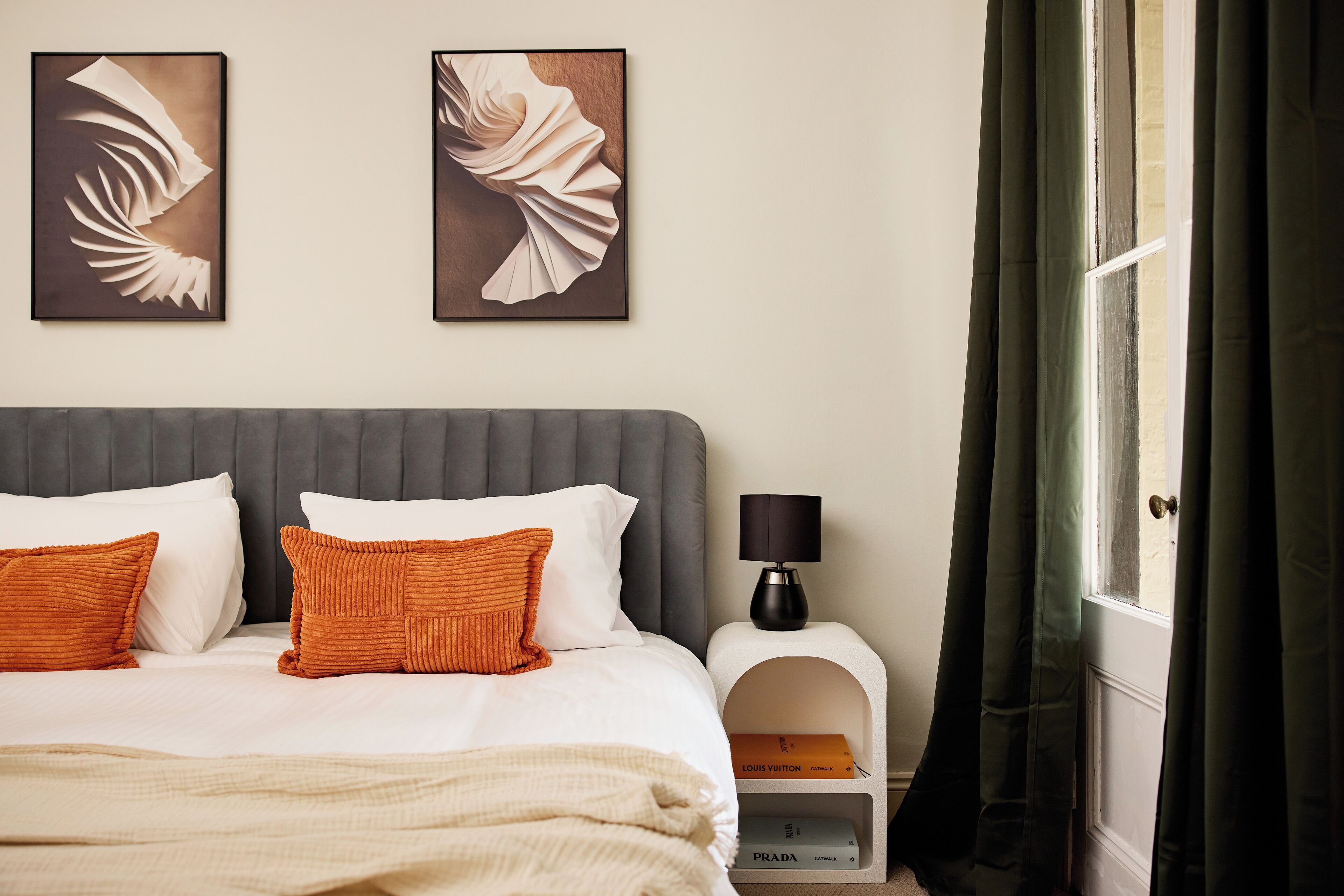 Een hotelkamer met een bed, kussens, een nachtkastje met een lamp en twee ingelijste kunstwerken aan de muur.