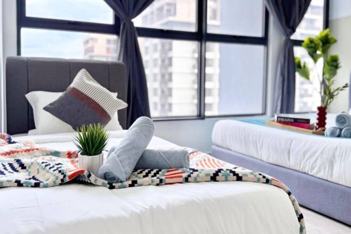 A modern bedroom with a bed, a bedside plant, and a view of buildings through large windows.