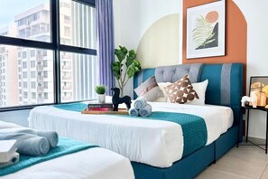 Hotellihuone, jossa on suuri ikkuna, sinisellä päätyosalla varustettu sänky, pieni huonekasvi ja seinällä oleva kehystetty kuva.