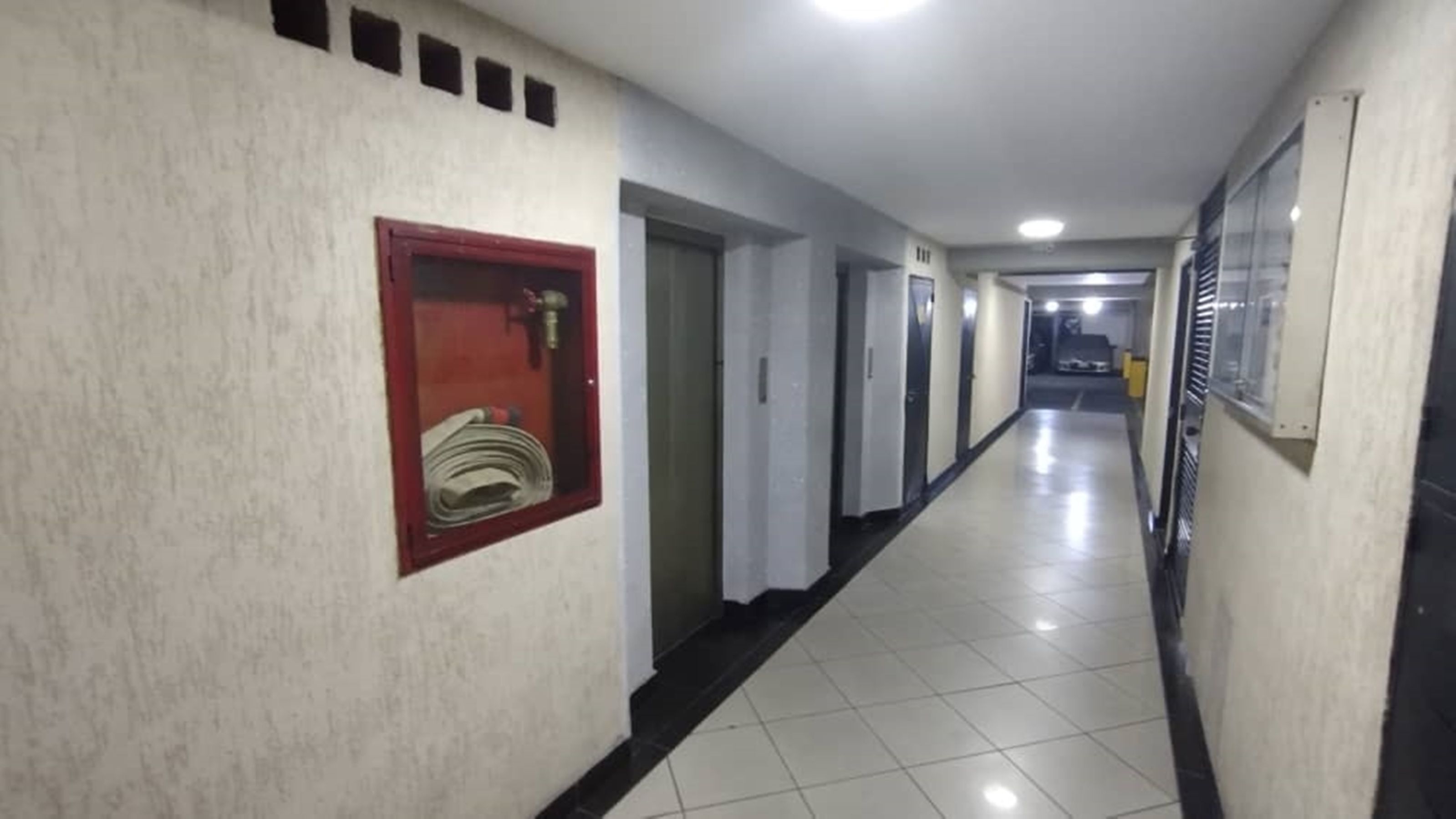 Un lungo e stretto corridoio con porte su entrambi i lati, un tubo antincendio montato sulla parete sinistra e una lampada a soffitto.