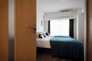 A hotel room with a bed, a window with curtains, an air conditioning unit, and a wooden door. - Tanimachikun Star Hotel Kujo 27 (Osaka)