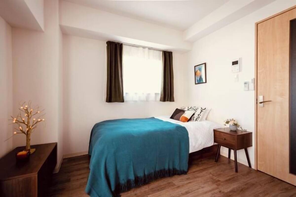 A bedroom with a bed, a bedside table, a nightstand, a window with curtains, a door, and a wall-mounted light switch.