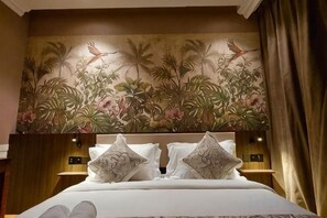 A hotel room with a bed, pillows, a headboard with a floral mural, and a nightstand with a lamp. - The Palm Suite at Lava Mansion (George Town)