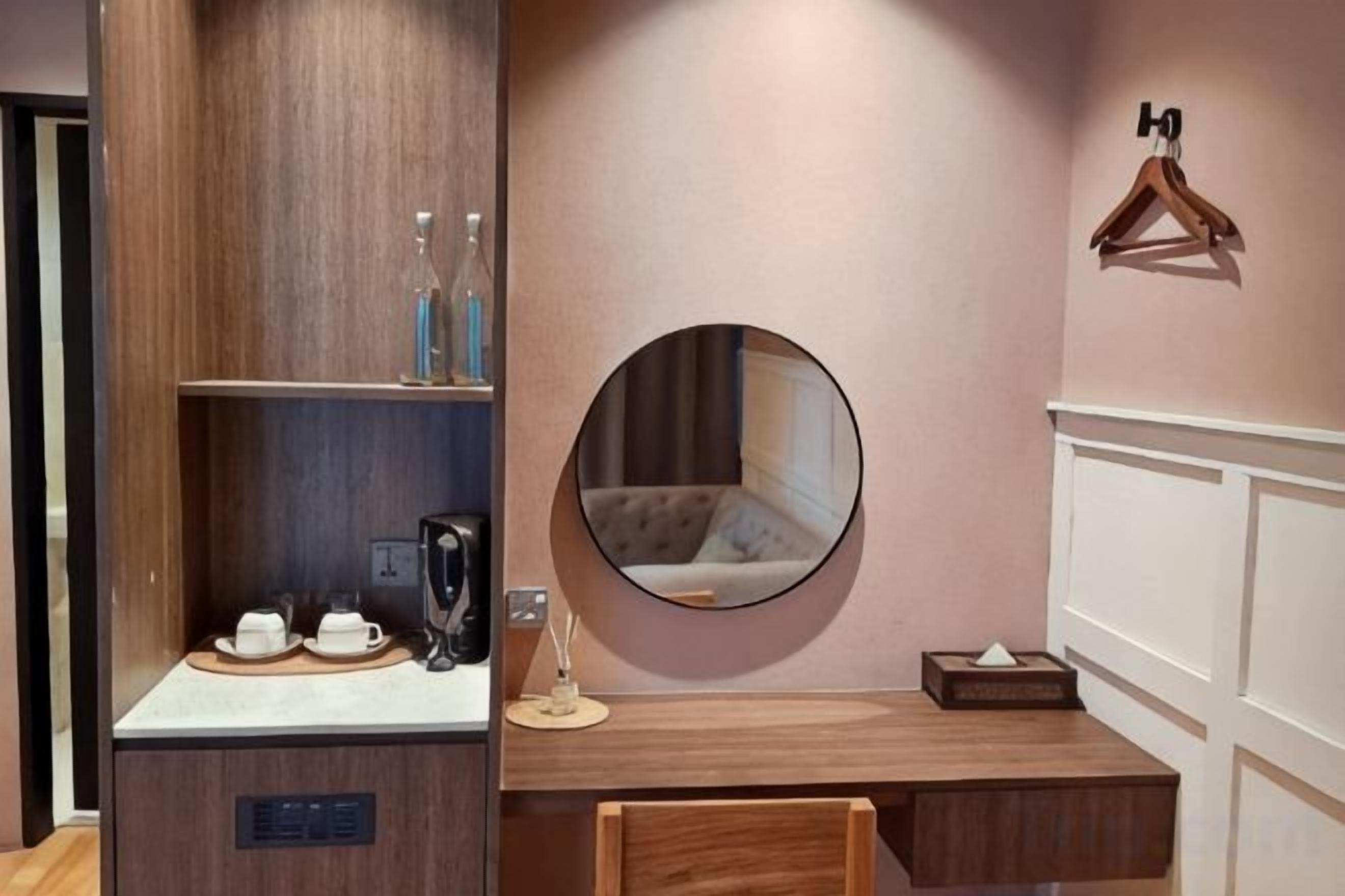 A hotel room with a wooden desk, a round mirror, a hanging coat rack, and a shelf with toiletries.
