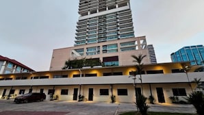 A multi-story hotel with a central courtyard, a parking area, and a tall building in the background. - The Palm Suite at Lava Mansion (George Town)