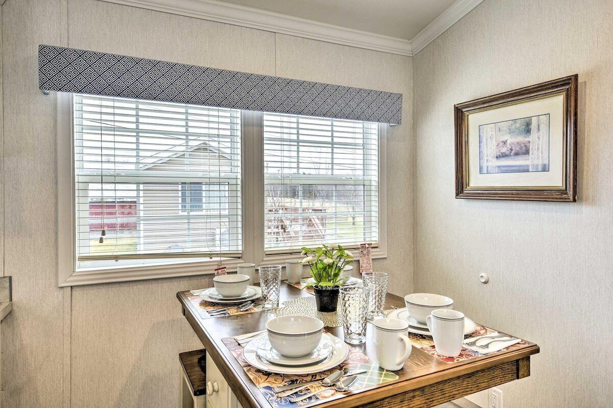 A dining area with a table set for four, two windows with blinds, a framed picture on the wall, and a potted plant on the windowsill.