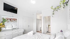 Une chambre à coucher moderne avec un grand lit, un téléviseur fixé au mur et une salle de bain visible à travers une porte ouverte.