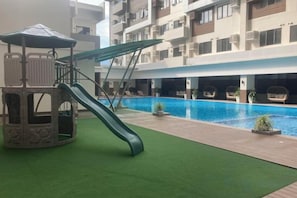 Un espace de jeux pour enfants avec un toboggan et une petite maison de jeu, une piscine et, au fond, un immeuble à plusieurs étages.