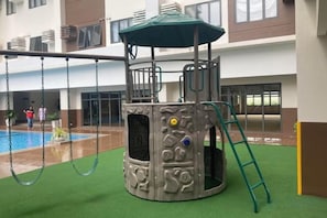 Un espace de jeux pour enfants avec une structure d’escalade, des balançoires et une piscine en arrière-plan.