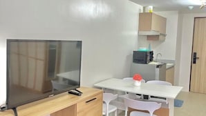 A compact room with a TV on a wooden stand, a small dining table with chairs, a microwave, and a cabinet. - By Trixia BE Apartments, Luxury Studio Apartments near IT Park (Cebu City)